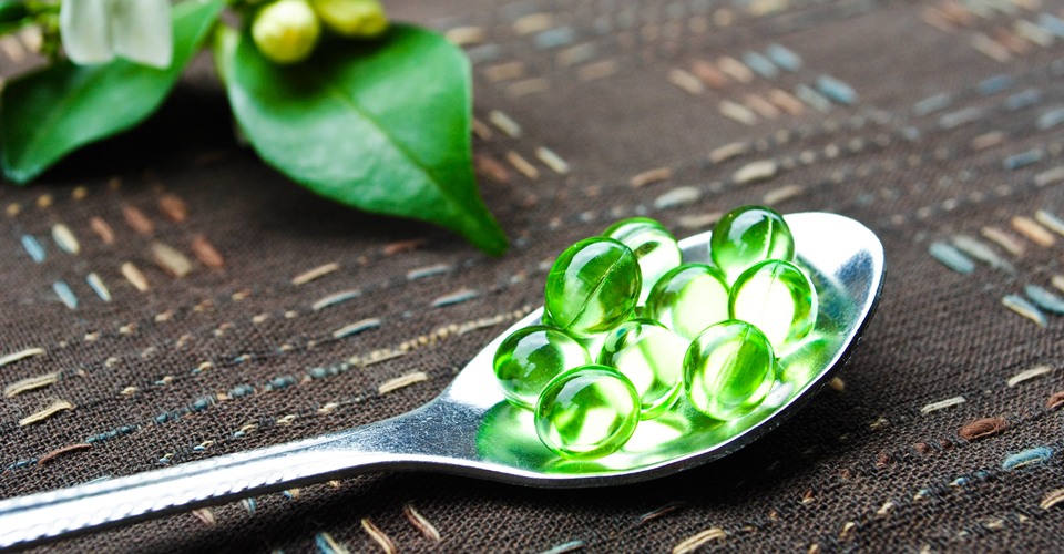 Green translucent gel capsules on a metal spoon with leaves in the background, dark textured cloth surface, calm natural mood